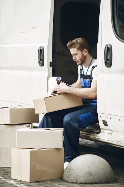 A man dressed in blue overalls is sitting on the open door sill of a white van, engaged in reviewing documents or inventory. Several cardboard boxes of varying sizes are placed on the ground nearby, suggesting the packing phase of a home relocation. The boxes are sealed with clear packing tape, some are open with flaps folded back, and others are closed, indicating organized packing and loading processes. The van’s interior is partially visible, showing more packing materials or boxes awaiting transport. The vehicle is parked on a paved surface, with a concrete bollard close to the van’s side, and the background suggests an outdoor setting during daylight. This scene illustrates professional furniture transport and moving logistics, as typical of home removals carried out by Uxbridge Man and Van, emphasizing careful handling, packaging, and planning involved in a household move.