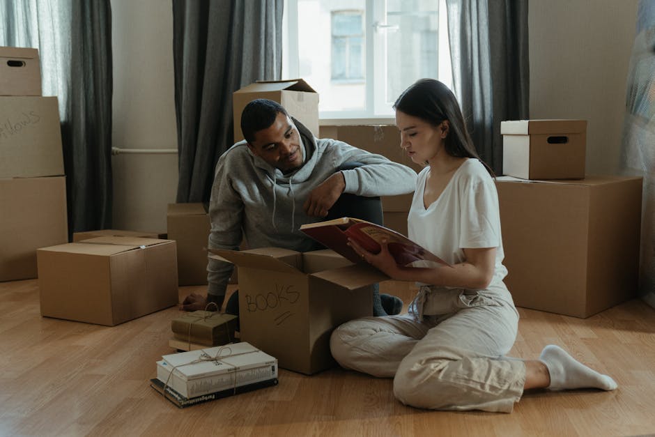 A man and a woman are inside a room filled with unpacked moving boxes, with some labeled 'books' on a cardboard box. The woman is sitting on the wooden floor, reading a magazine, while the man is leaning over a partially opened cardboard box, looking at her. Around them, there are several closed cardboard boxes of various sizes stacked against the wall and near a window with natural light coming in, suggesting an ongoing home relocation process. The floor is clear except for the boxes and a few packages, and the setting indicates furniture transport and packing activities typical during house removals. Uxbridge Man and Van uses this scene to illustrate the logistics involved in furniture transport and packing for house moves, supporting services like packing and moving for students at Brunel University.