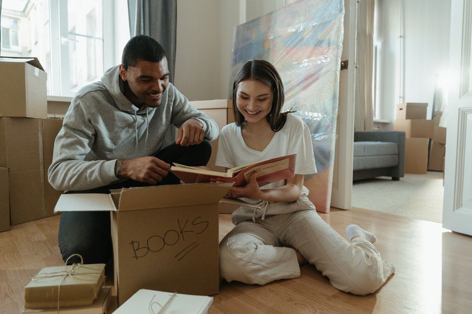 A young man and woman sit on a light wooden floor inside a room with natural daylight coming through a large window, surrounded by packing boxes and stacked cardboard cartons. The man, dressed in a grey hoodie, is holding an open book while smiling at the woman, who is wearing a white t-shirt and beige joggers. The woman is holding a smaller book and appears engaged in reading or conversation. There is a cardboard box labeled 'BOOKS' in front of them, with some wrapped packages and notebooks nearby. In the background, a partially covered large mirror or artwork is leaning against the wall, with a grey sofa and additional cardboard boxes placed along the walls, indicating a house removal or packing process. The scene captures the packing and home relocation activities associated with furniture transport and moving logistics, as handled by Uxbridge Man and Van, supporting a smooth transition during house removals or student relocations at Brunel University in Uxbridge.