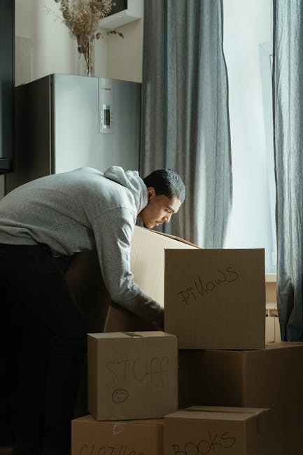 A man and a woman are inside a room filled with unpacked moving boxes, with some labeled 'books' on a cardboard box. The woman is sitting on the wooden floor, reading a magazine, while the man is leaning over a partially opened cardboard box, looking at her. Around them, there are several closed cardboard boxes of various sizes stacked against the wall and near a window with natural light coming in, suggesting an ongoing home relocation process. The floor is clear except for the boxes and a few packages, and the setting indicates furniture transport and packing activities typical during house removals. Uxbridge Man and Van uses this scene to illustrate the logistics involved in furniture transport and packing for house moves, supporting services like packing and moving for students at Brunel University.